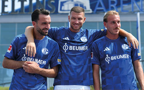 Drei Erwachsene in blauen Fu balltrikots mit dem Schriftzug Beumer stehen eng beieinander vor einem Stadion und l cheln. Beschreibung von KI generiert