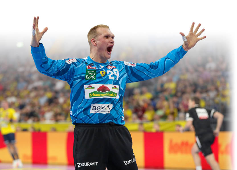 Ein jubelnder Spieler in blauem Trikot und schwarzer Hose steht mit weit ge ffneten Armen und offenem Mund in einer Sporthalle vor einem unscharfen Publikum. Beschreibung von KI generiert