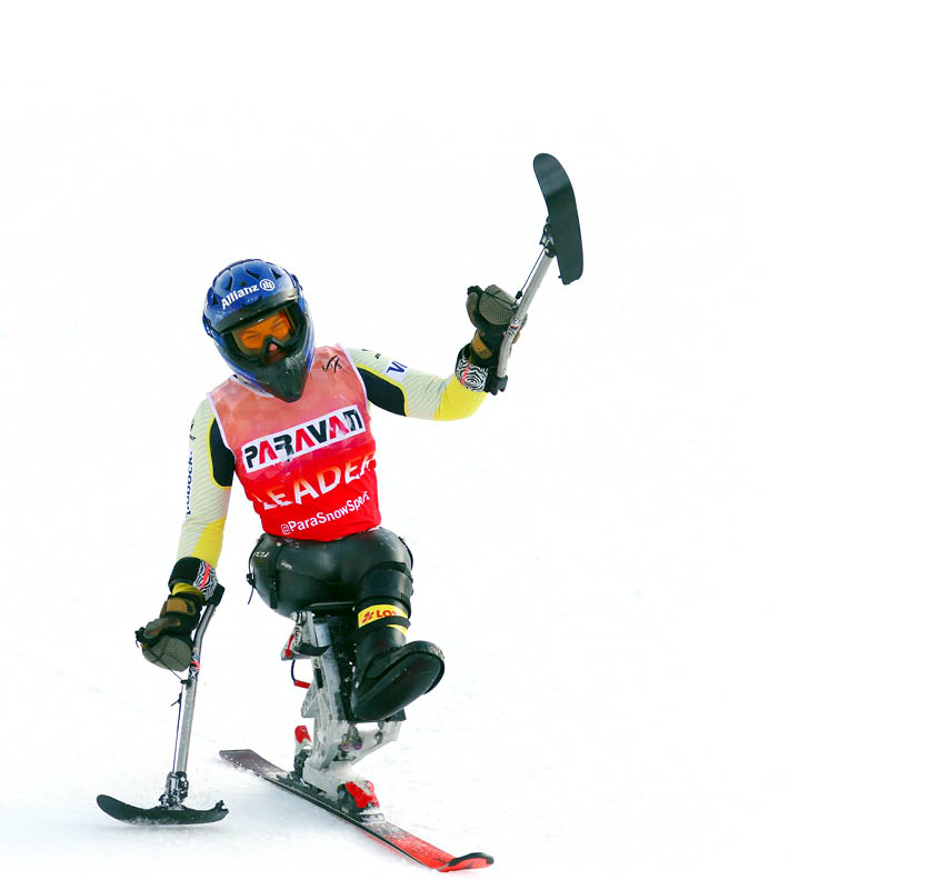 Eine Person mit Helm und Schutzkleidung f hrt mit einem Monoski auf Schnee und hebt einen Stock in die Luft. Beschreibung von KI generiert